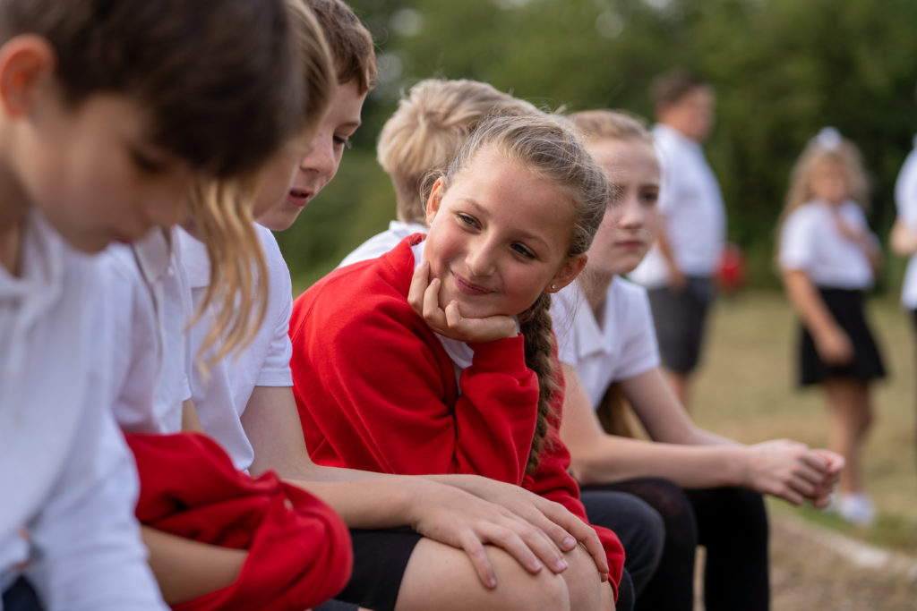 Six older pupils are shown sitting together on a bench outdoors. One girl is smiling at the others with her chin resting on the palm of her right hand.